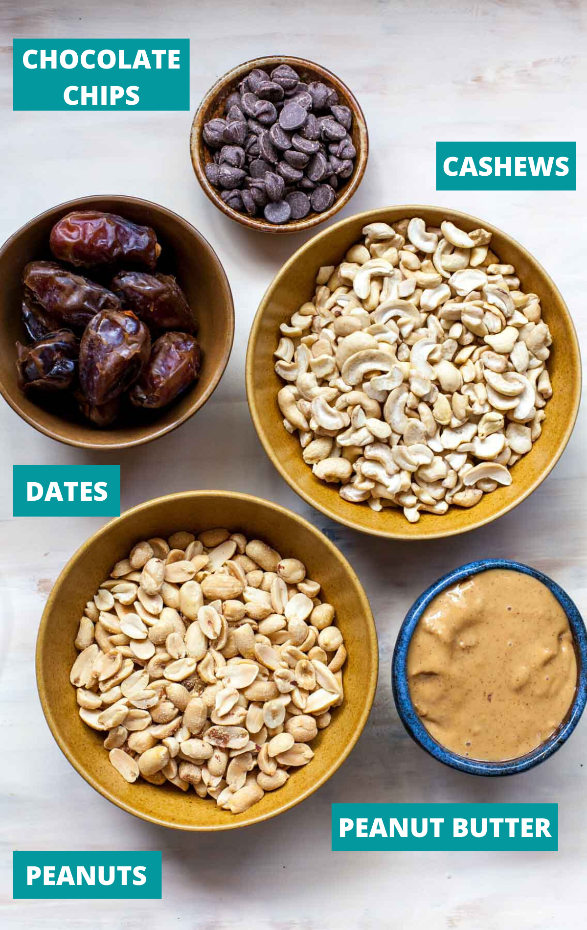 Nuts, dates, chocolate chips, and peanut butter in separate bowls on a white board