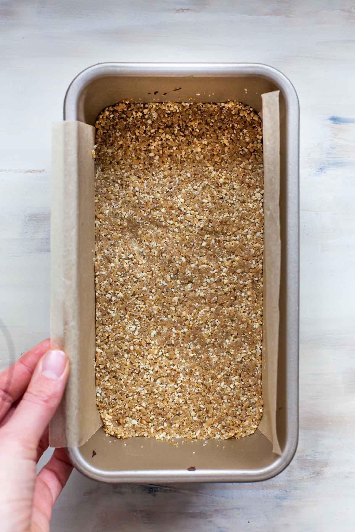 Shortbread cookie crust being pressed into a loaf pan covered in parchment paper