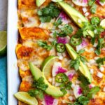 Zucchini and quinoa enchiladas styled in a white pan topped with avocado, radish slices, and cilantro.