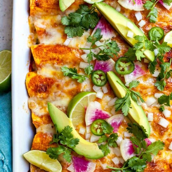 Zucchini and quinoa enchiladas styled in a white pan topped with avocado, radish slices, and cilantro.