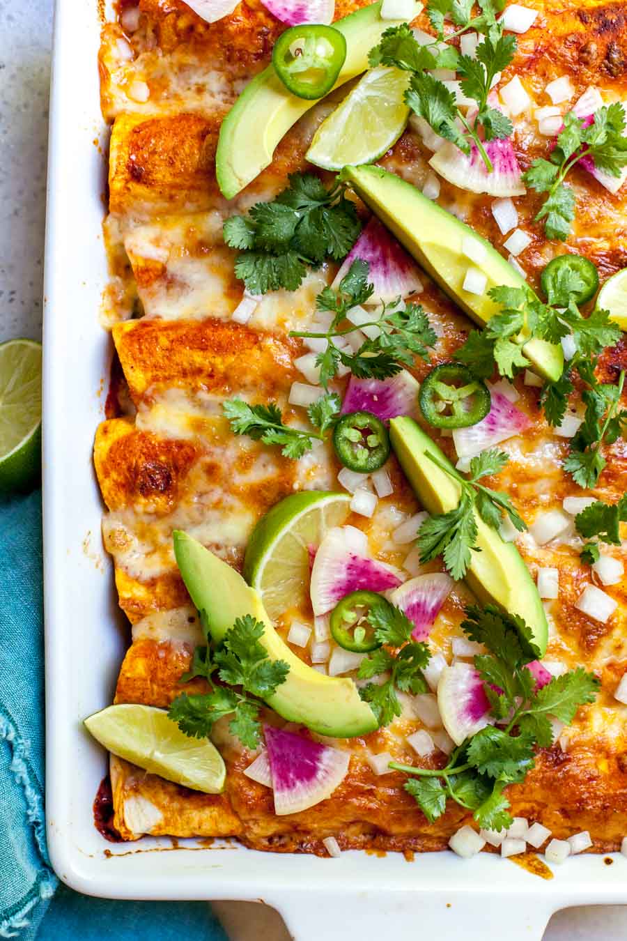 Zucchini and quinoa enchiladas styled in a white pan topped with avocado, radish slices, and cilantro.
