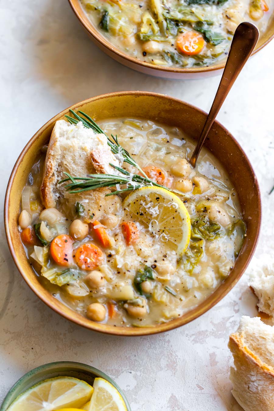Bowl of white bean soup topped with a lemon slice, fresh rosemary, and crusty bread
