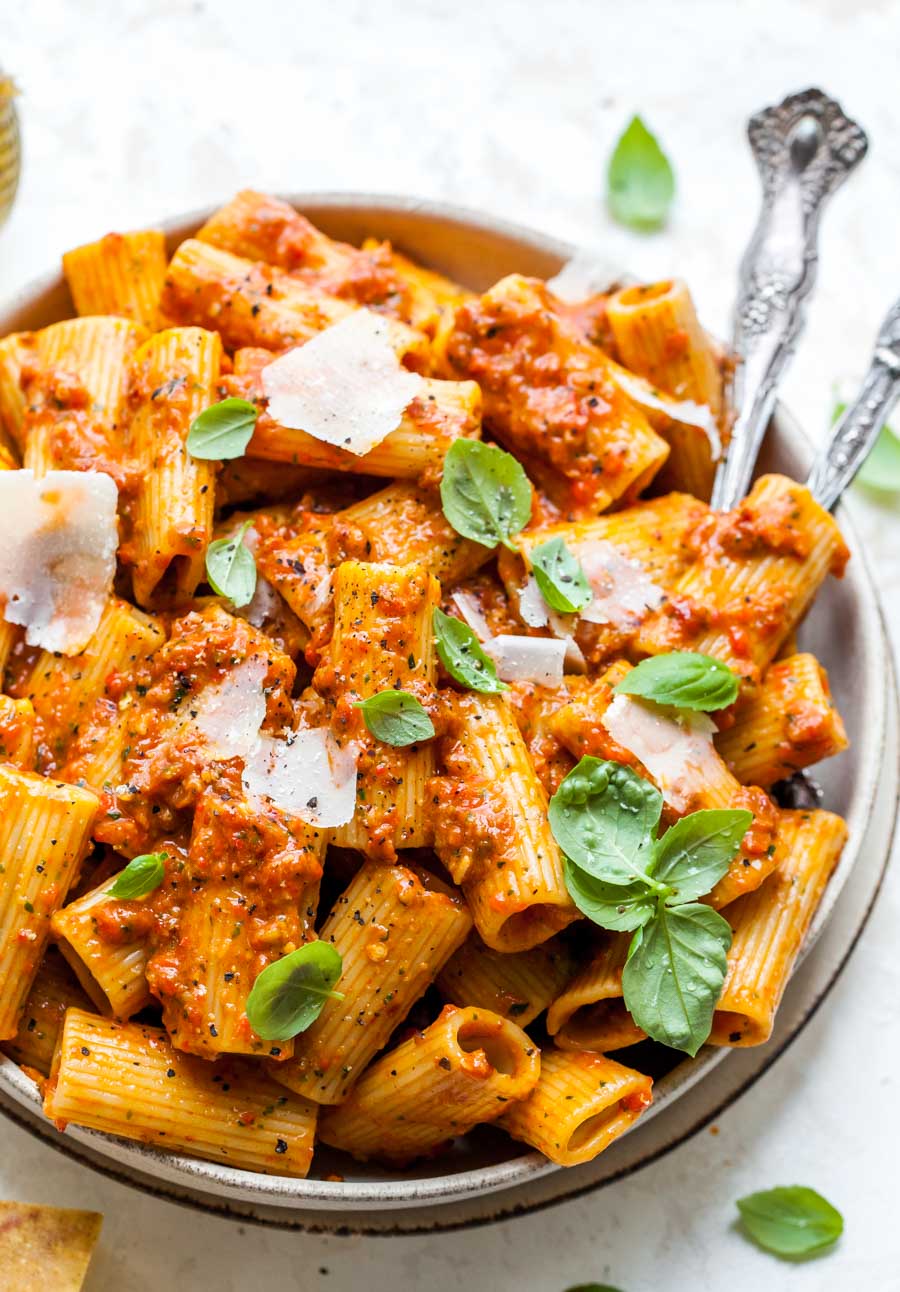 Rigatoni tossed with red pesto in a serving bowl with serving utensils resting on the right side.