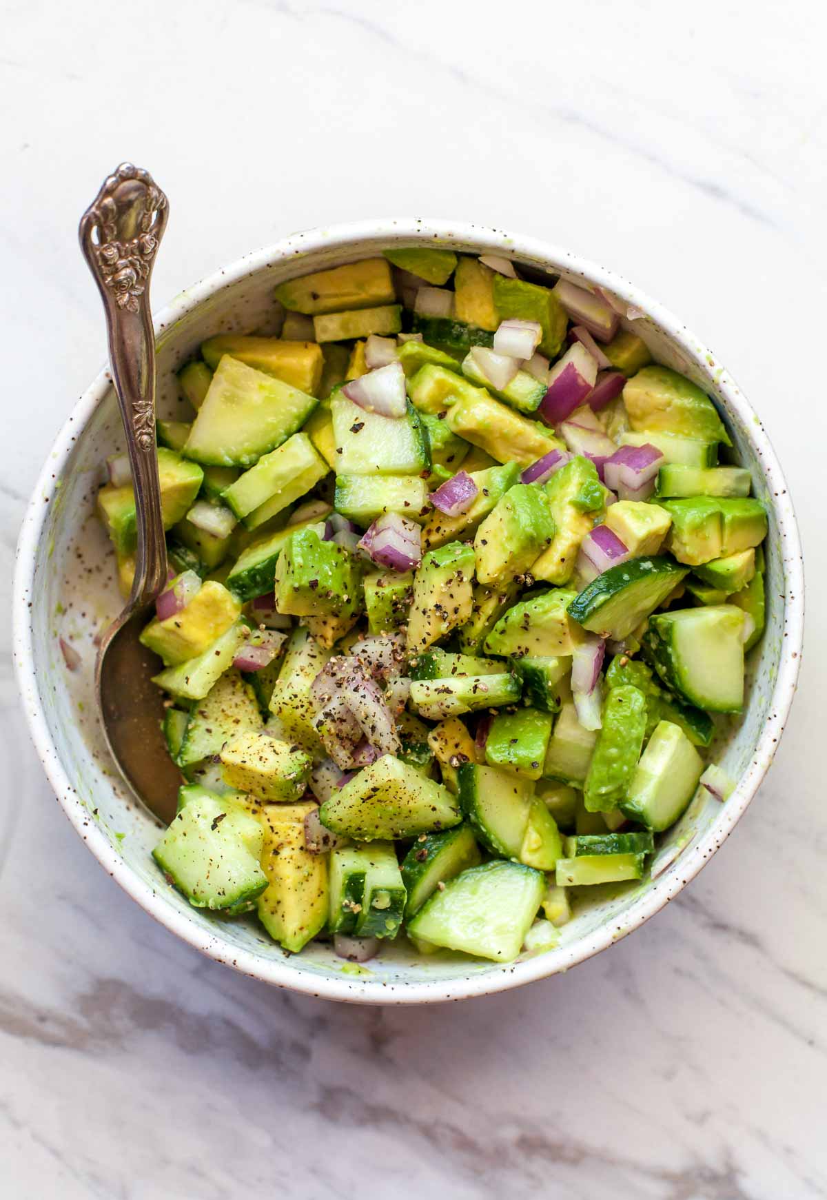 Chopped avocado, cucumber, and red onion in a white bowl with a spoon resting on the left side