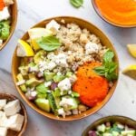 Quinoa, chickpeas, and cucumber salad in a gold bowl with red pepper sauce spooned overtop