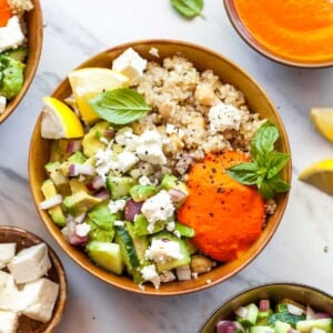 Quinoa, chickpeas, and cucumber salad in a gold bowl with red pepper sauce spooned overtop