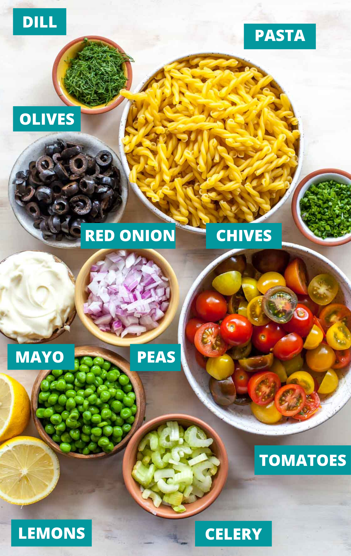 Dry pasta, vegetables, and herbs in separate small bowls on a tan surface with blue ingredient labels
