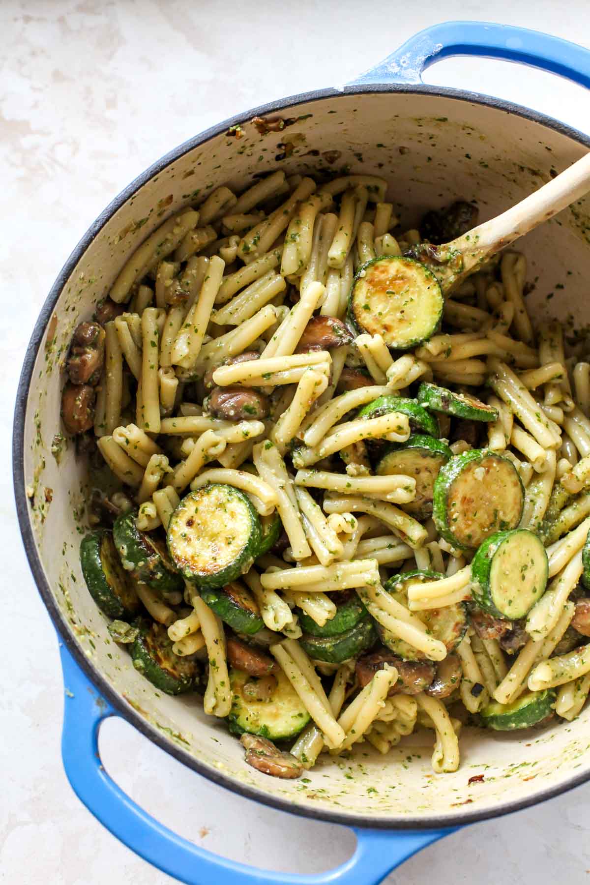 Pasta mixed with pesto and zucchini in a large blue pot with a wooden spoon.