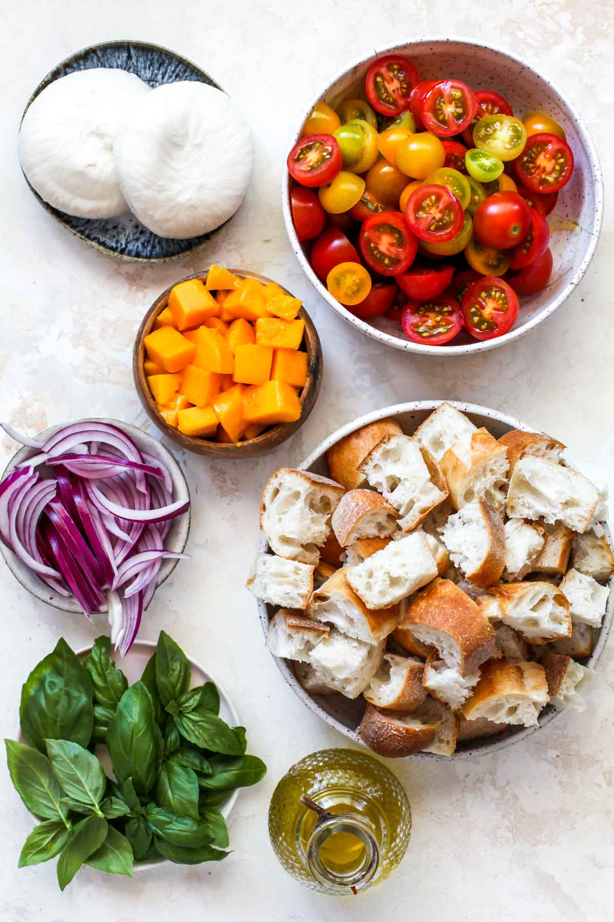 Tomatoes, bread, basil, mango, and red onion in separate bowls arranged on a white surface.