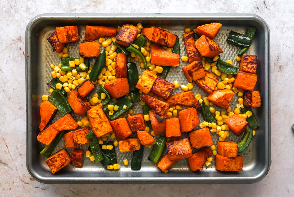 Corn, squash, and peppers being mixed together on a baking sheet