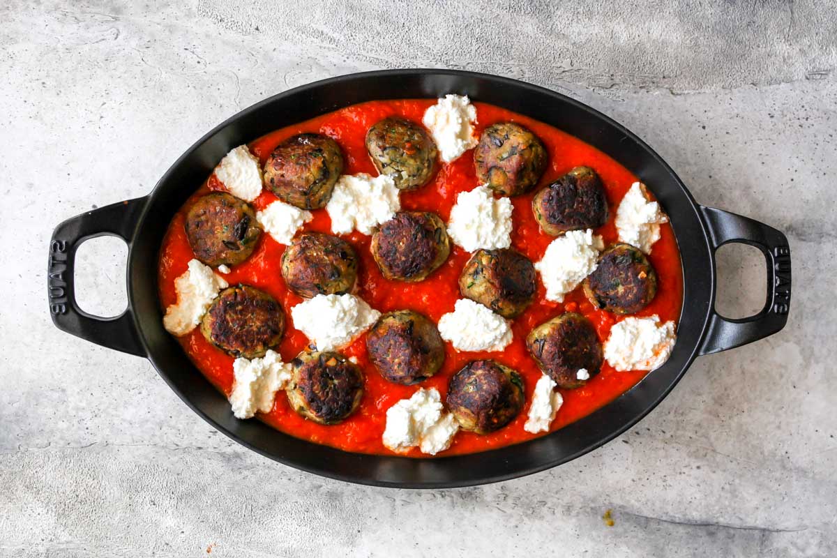 Marinara sauce, dollops of ricotta cheese, and eggplant meatballs arranged in an oval baking dish before baking.