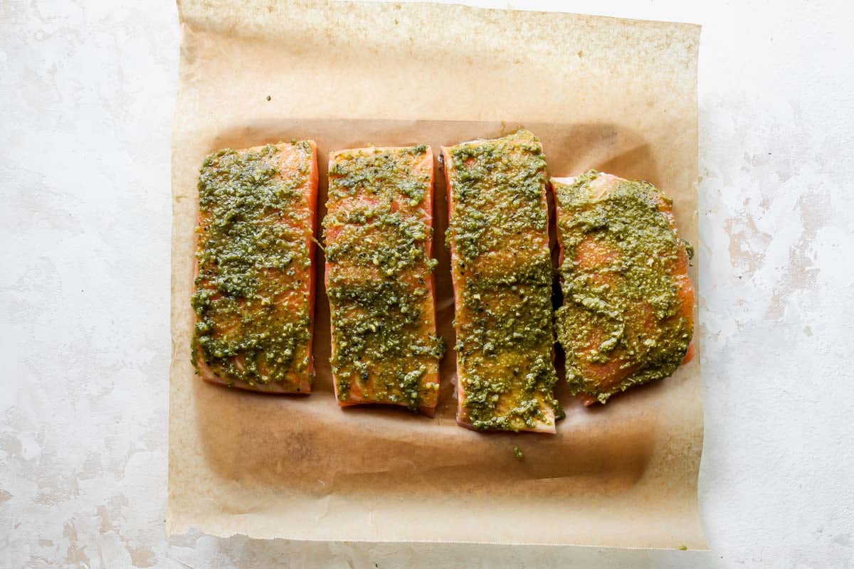Four salmon fillets on a cutting board being rubbed with pesto