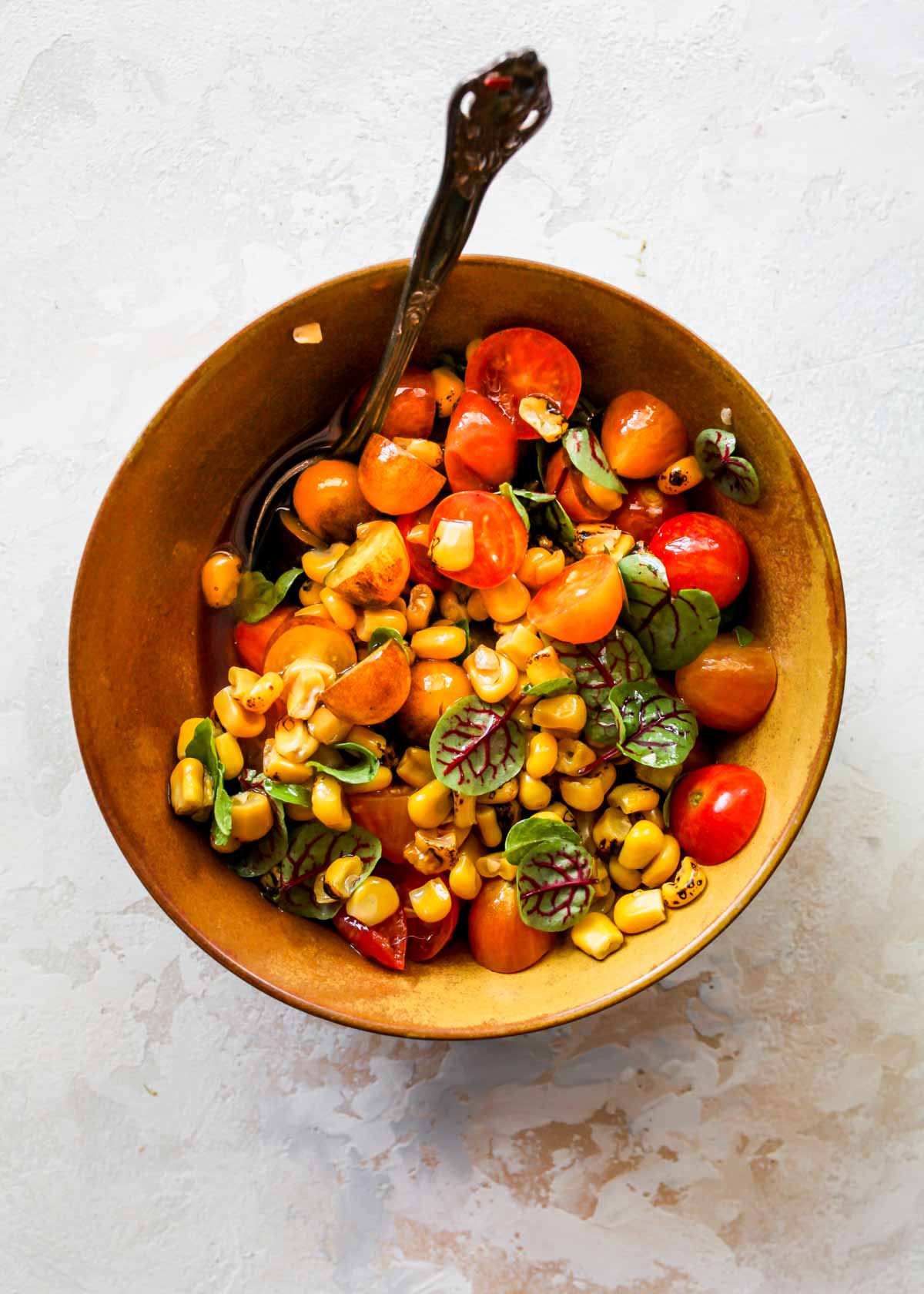 Cherry tomatoes, corn, basil, vinegar, and olive oil being stirred in a bowl