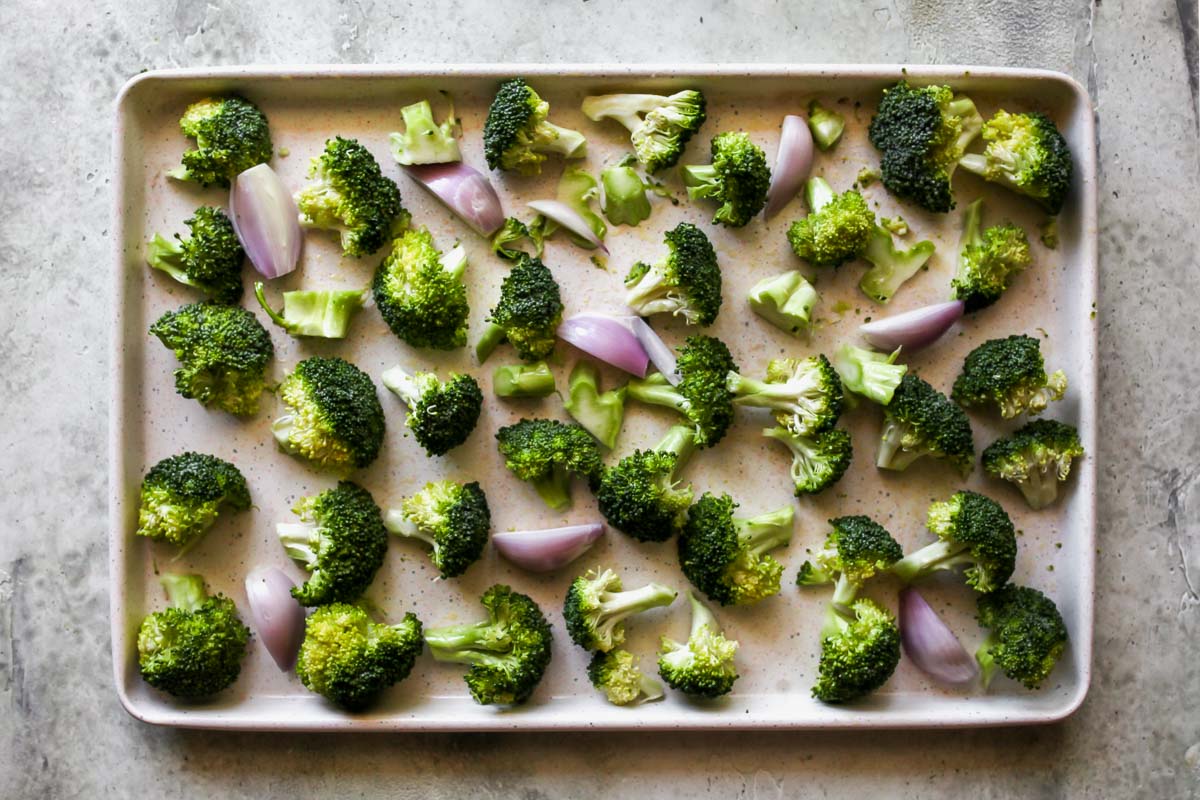 Broccoli florets and shallots arranged on a white baking sheet.