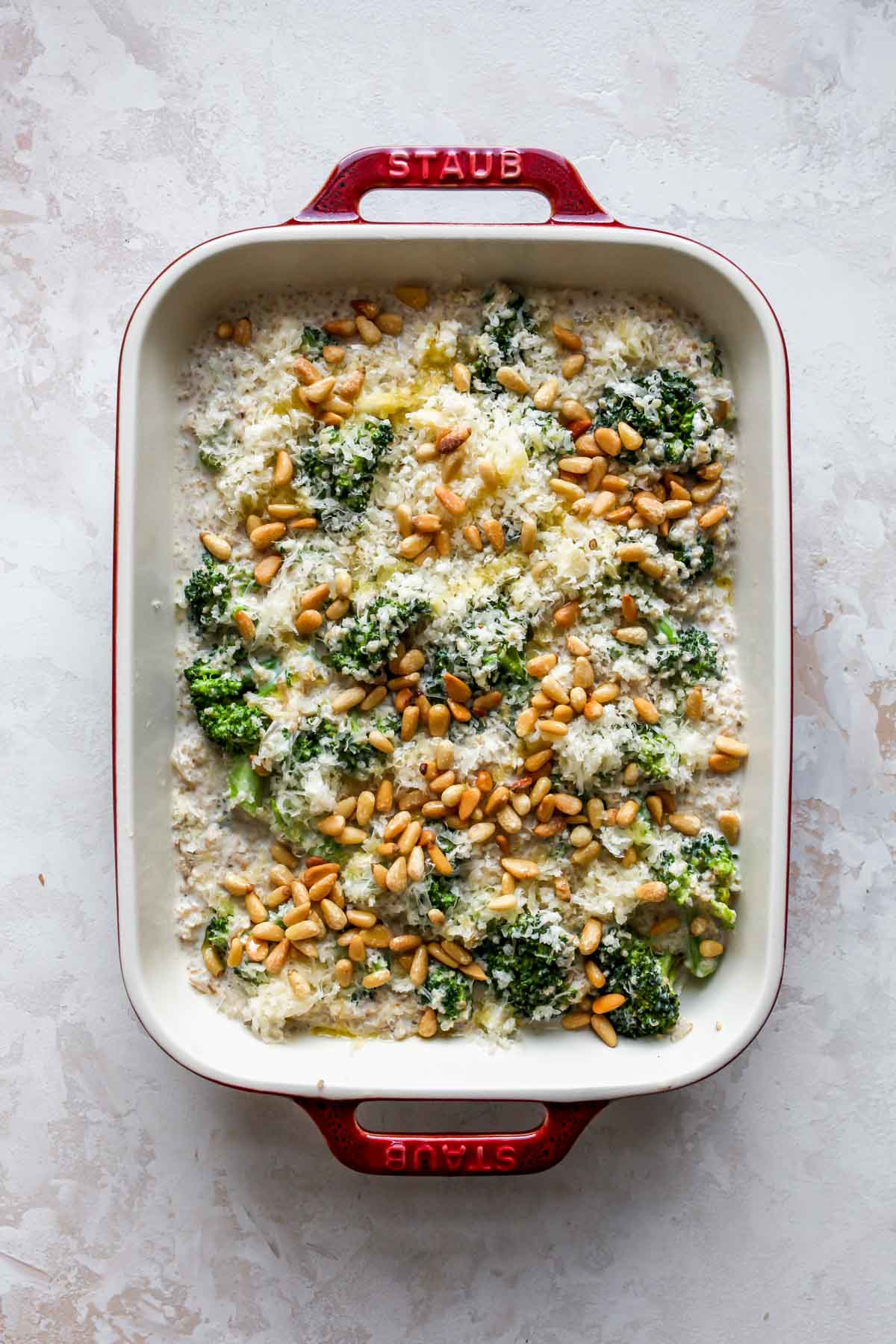 Quinoa and broccoli mixture in a red baking dish topped with pine nuts before baking
