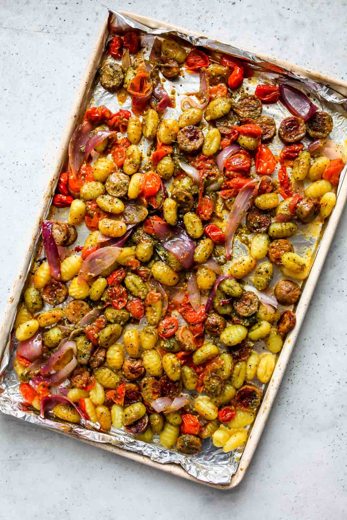 Pesto being mixed into pasta and veggie mixture on a sheet pan