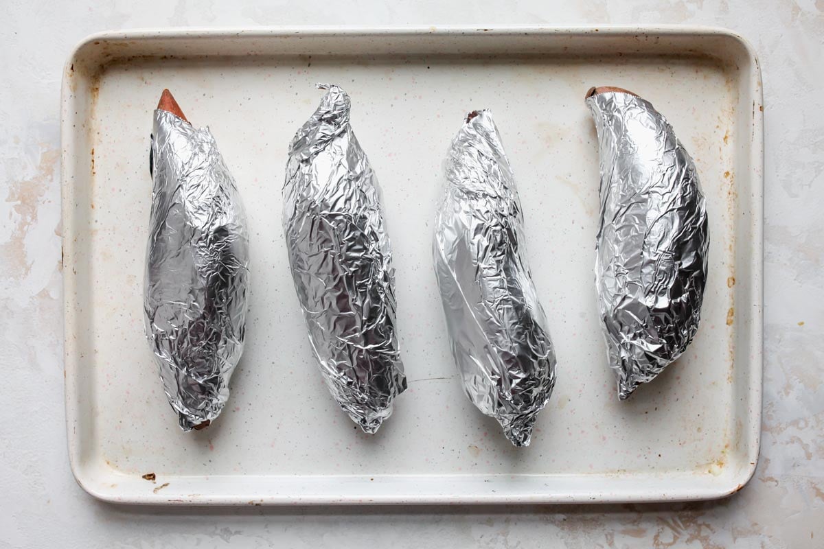 Sweet potatoes wrapped in foil and being roasted on a sheet pan
