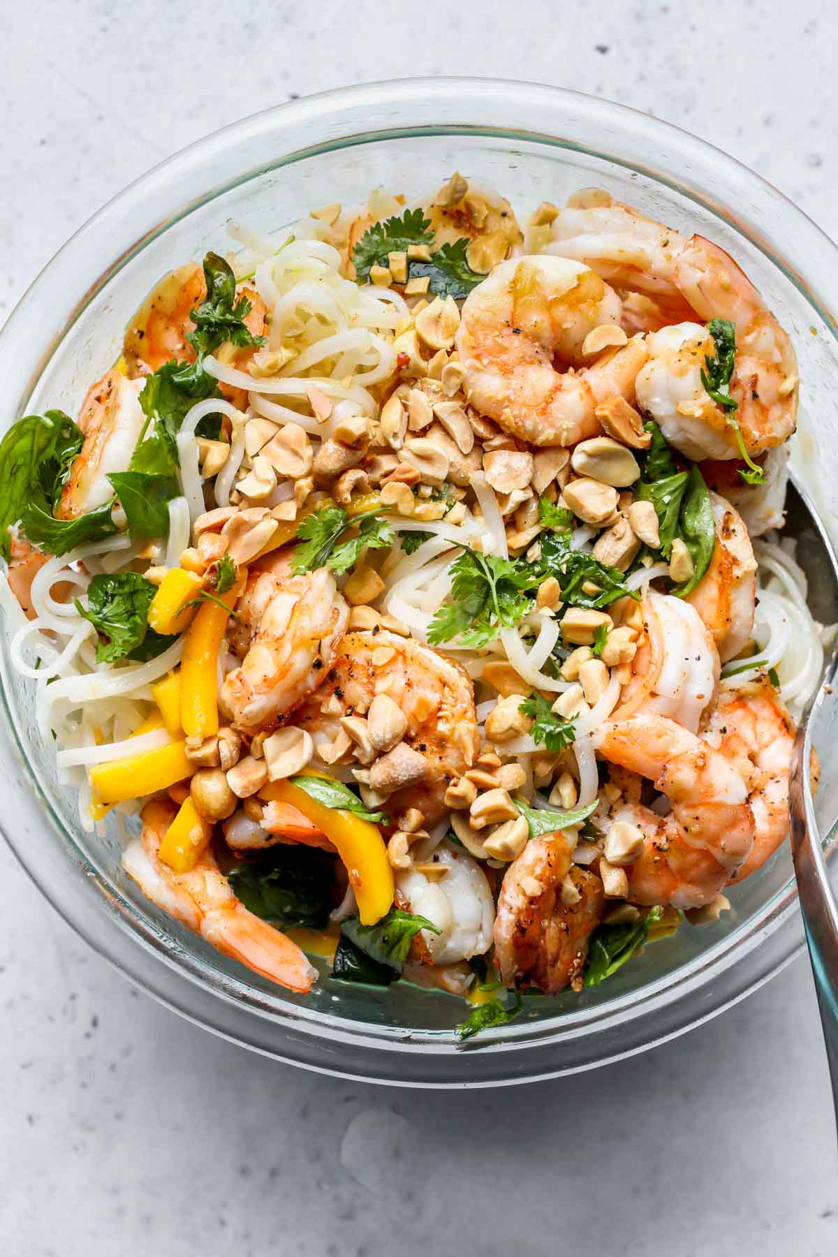 Noodles being tossed with shrimp, mango, herbs, and peanuts in a glass mixing bowl