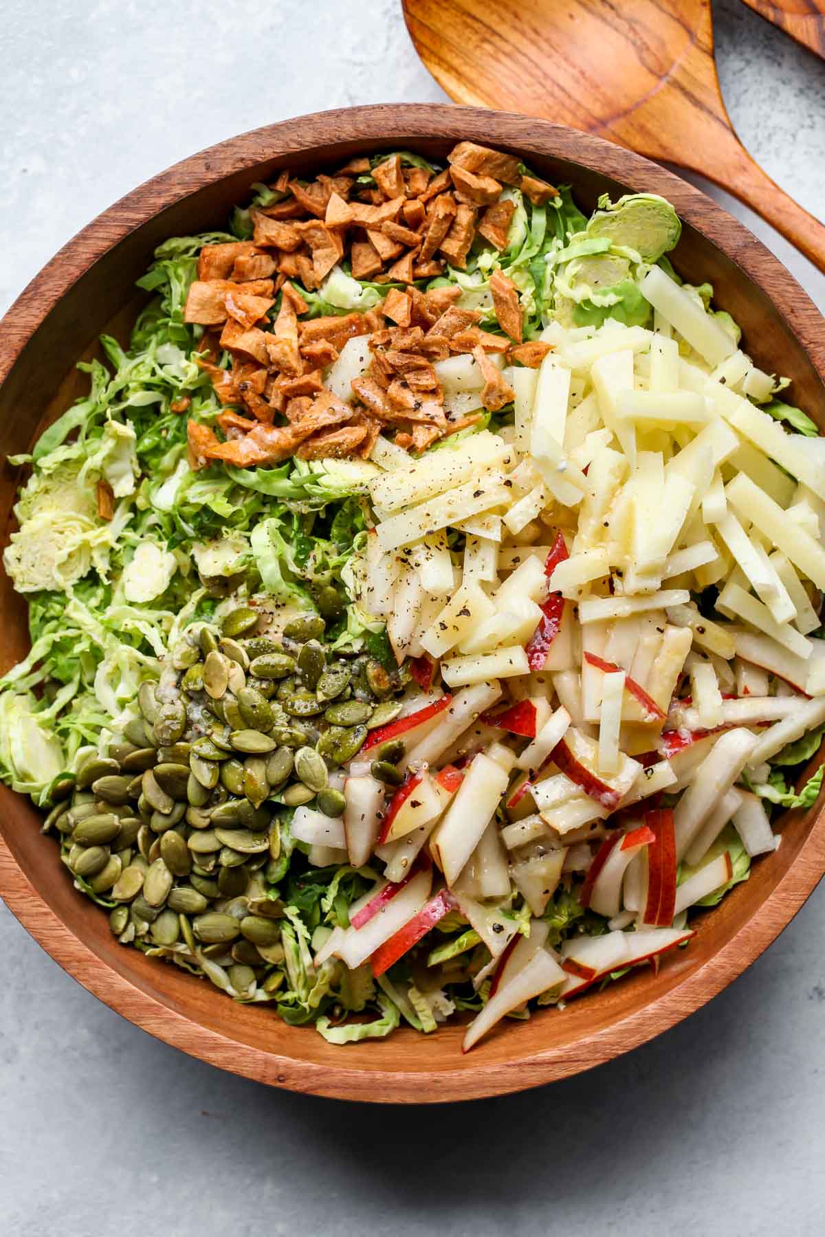 Shaved brussels, pear, dried apple, peptias, and cheese being added to a wooden mixing bowl