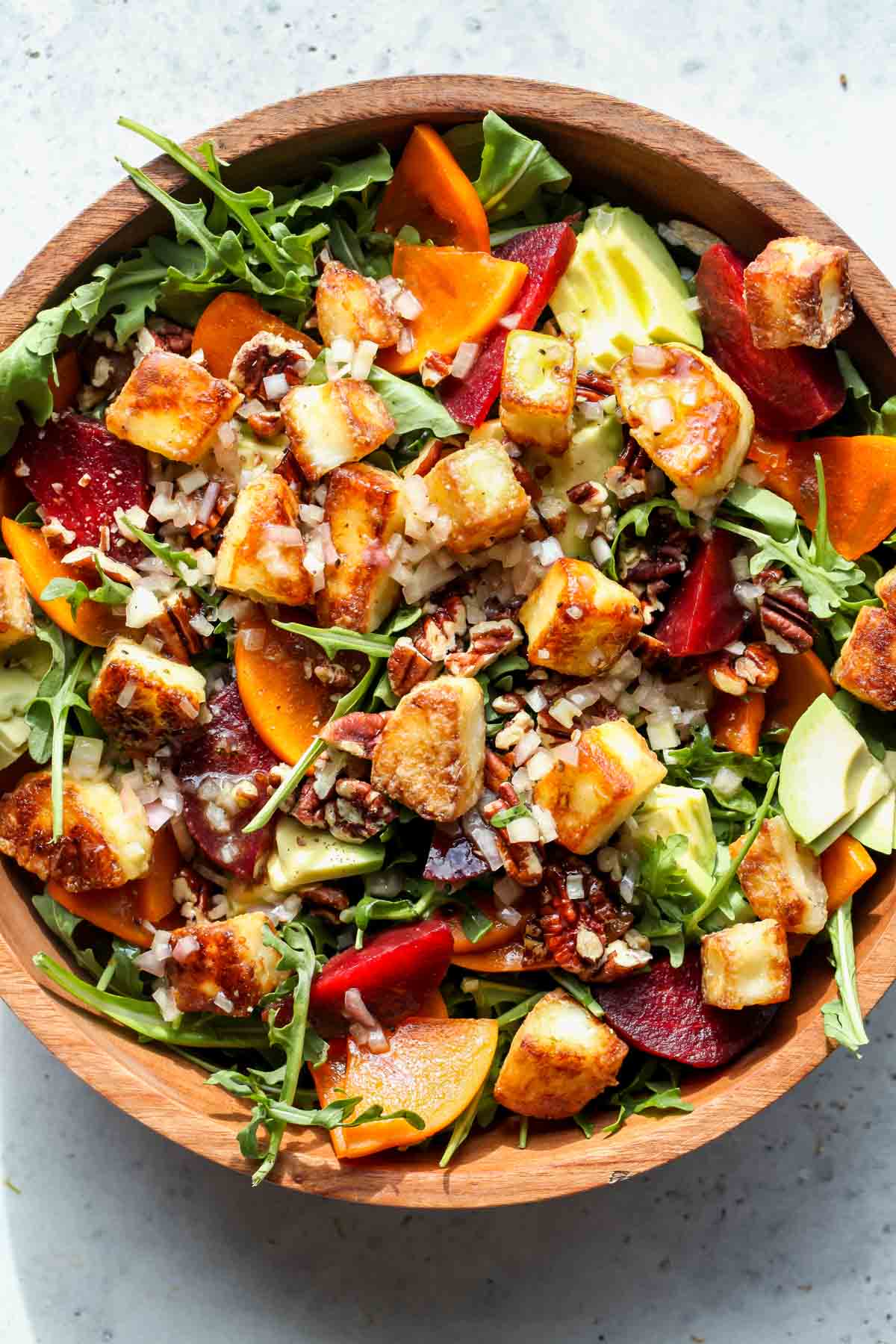 Arugula, persimmons, sliced beets, avocado, pecans, and halloumi being assembled in a large wood serving bowl