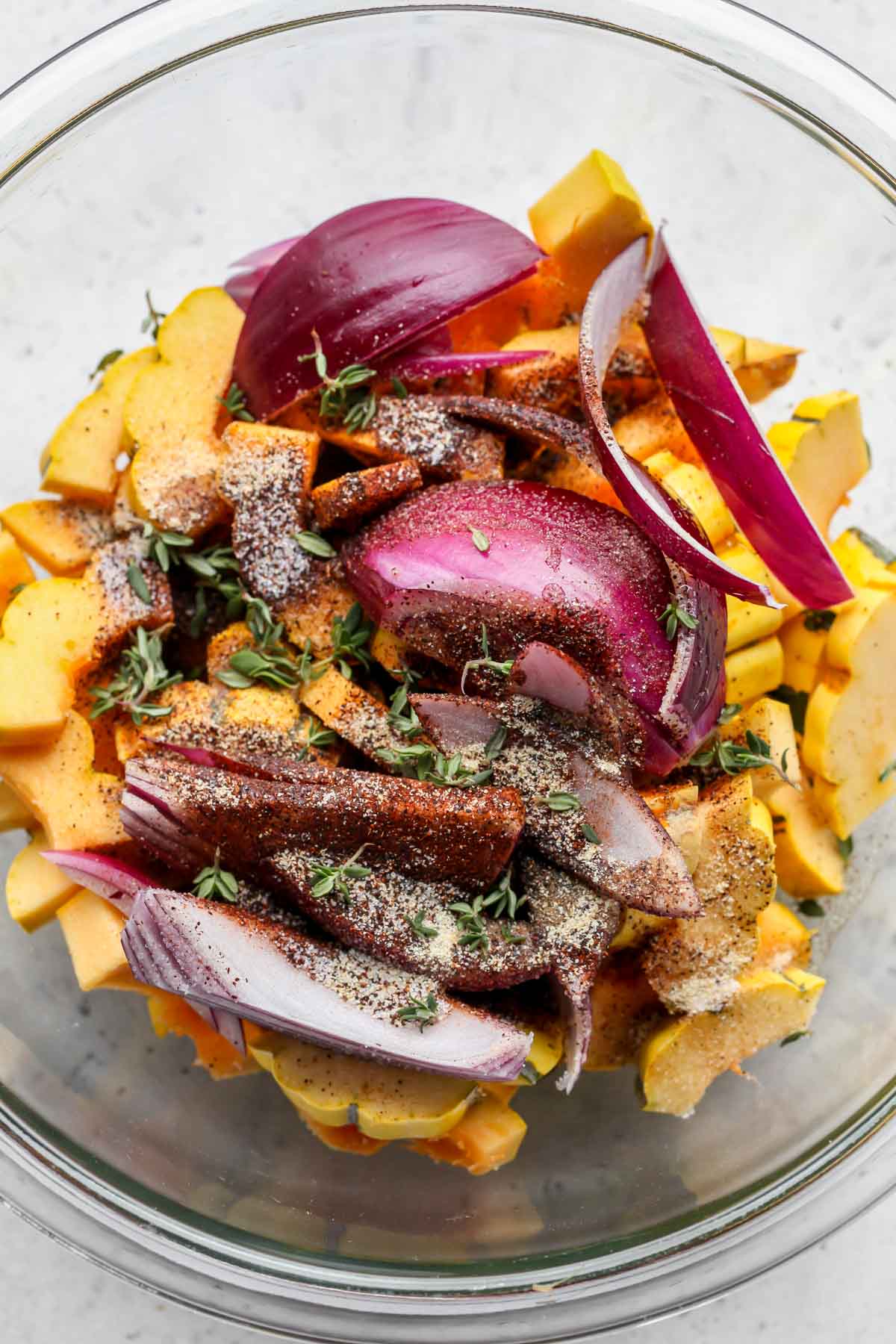 Sliced delicata squash and red onion in a large bowl being tossed in oil, spices, and herbs