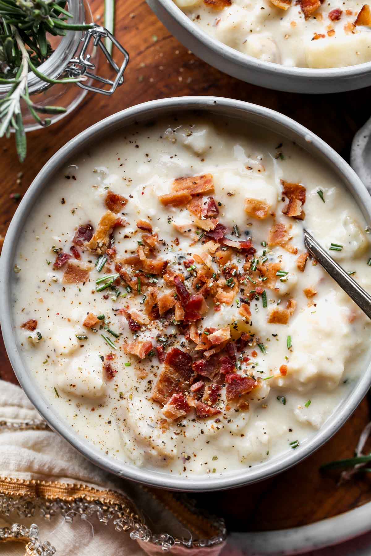 Spoon resting in a bowl of crock pot potato soup garnished with bacon and rosemary
