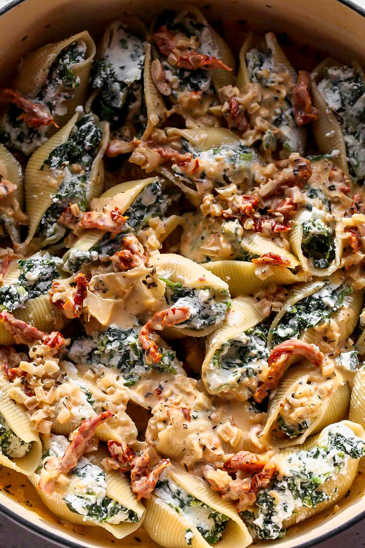 Sun-dried tomato sauce being poured over stuffed shells in a baking dish
