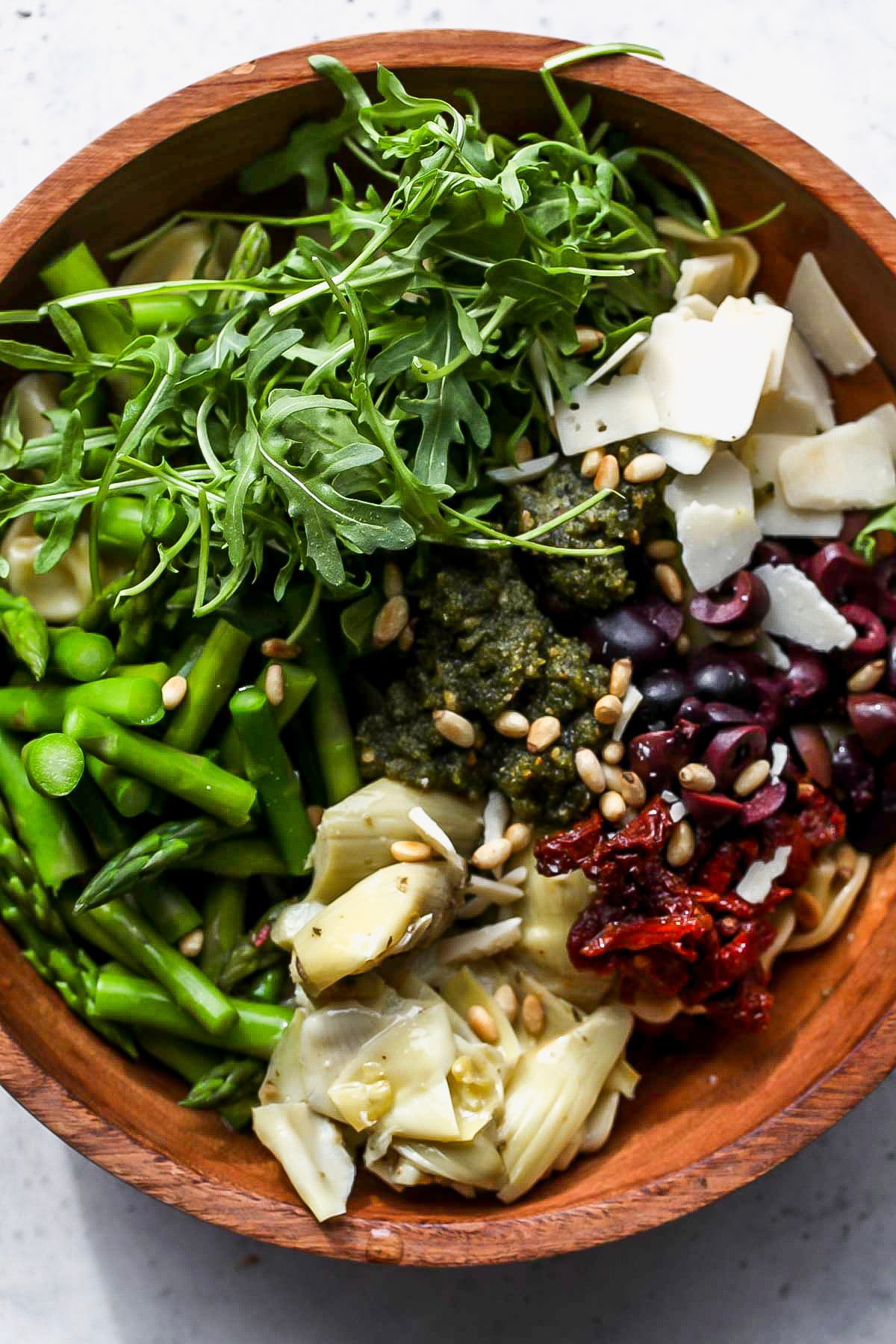 Tortellini, asparagus, arugula, pesto, artichokes, and sun-dried tomatoes being added to a wooden bowl