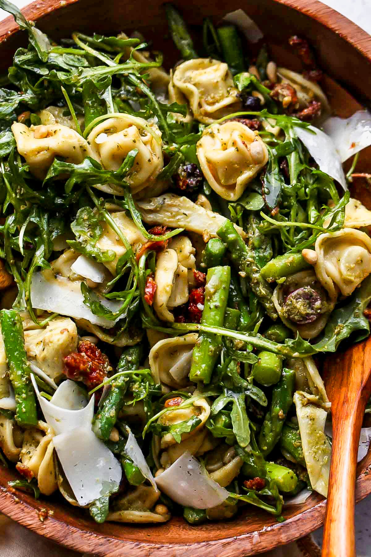 Tortellini pasta salad being tossed with wooden utensils in a bowl.