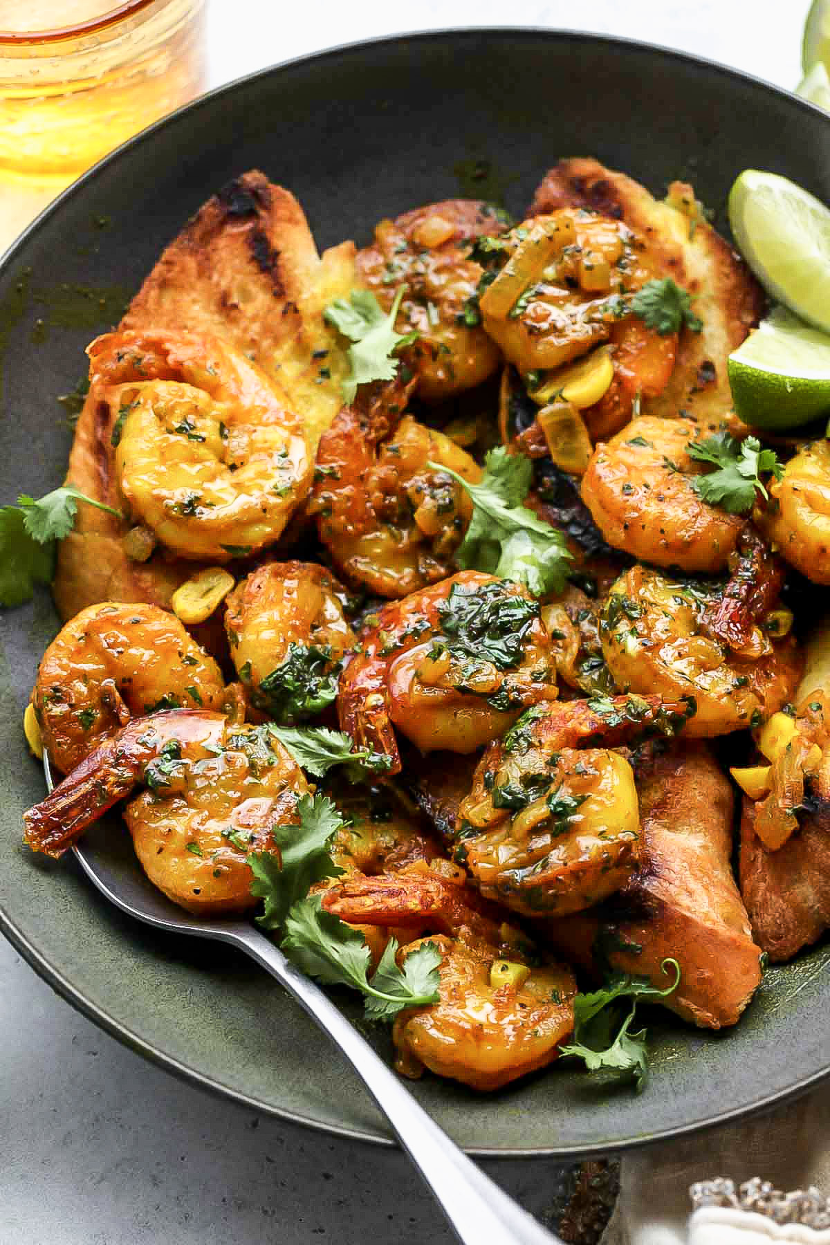 A serving spoon digging into a bowl of sautéed shrimp in turmeric sauce with lime wedges.