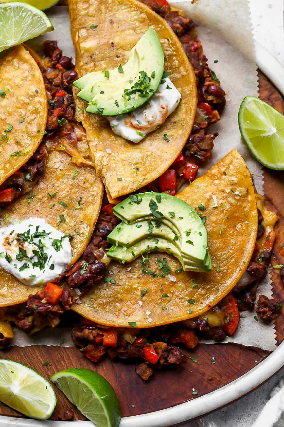 Crispy black bean tacos topped with sour cream, avocado, and cilantro arranged on a wooden serving platter.