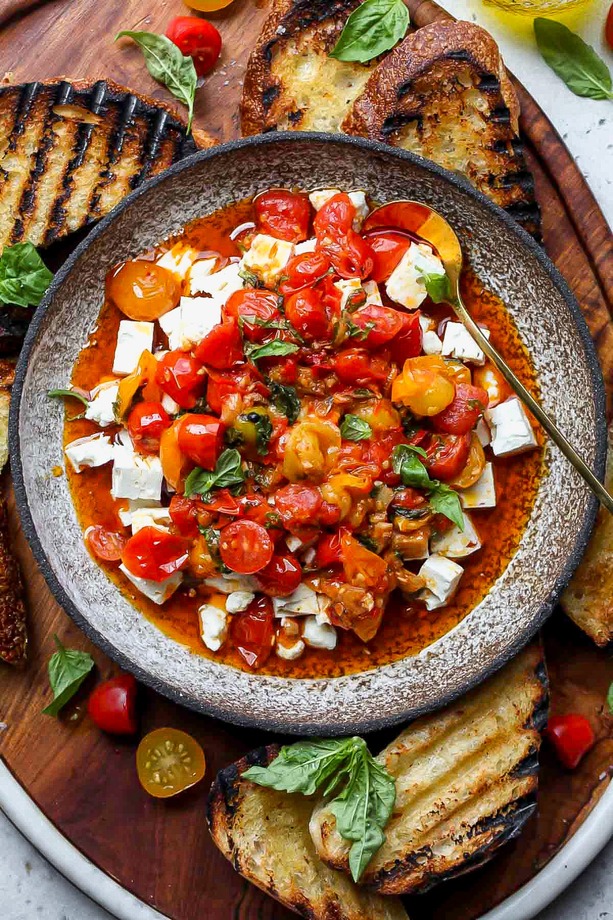 Spicy tomato marinated feta in a serving bowl surrounded by grilled bread.