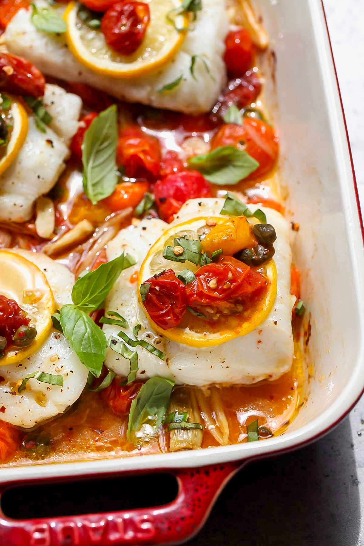 Mediterranean baked cod in a red baking dish topped with fresh basil and lemon slices.