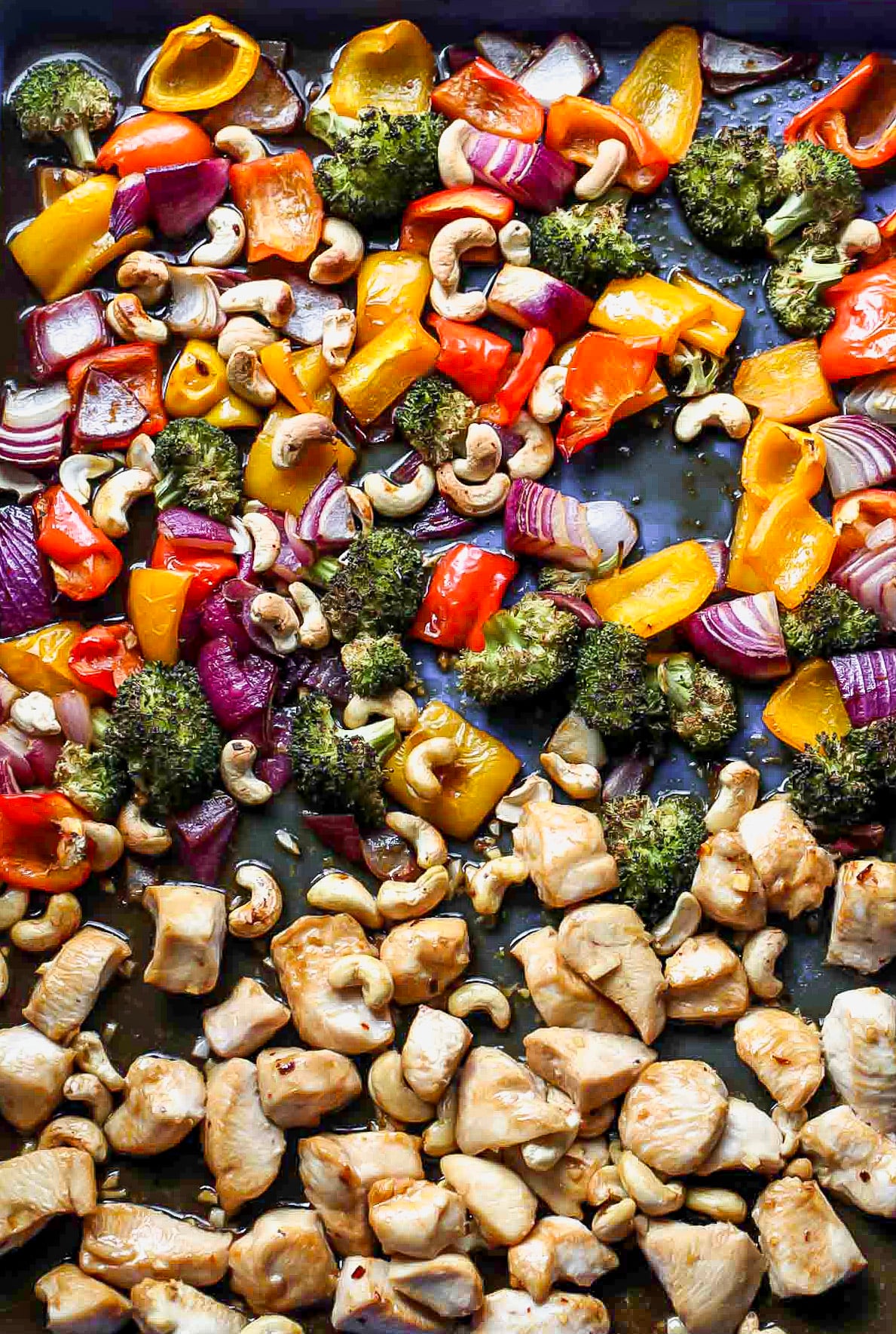 Assorted vegetables, chicken, and cashews on a large baking sheet after being roasted.