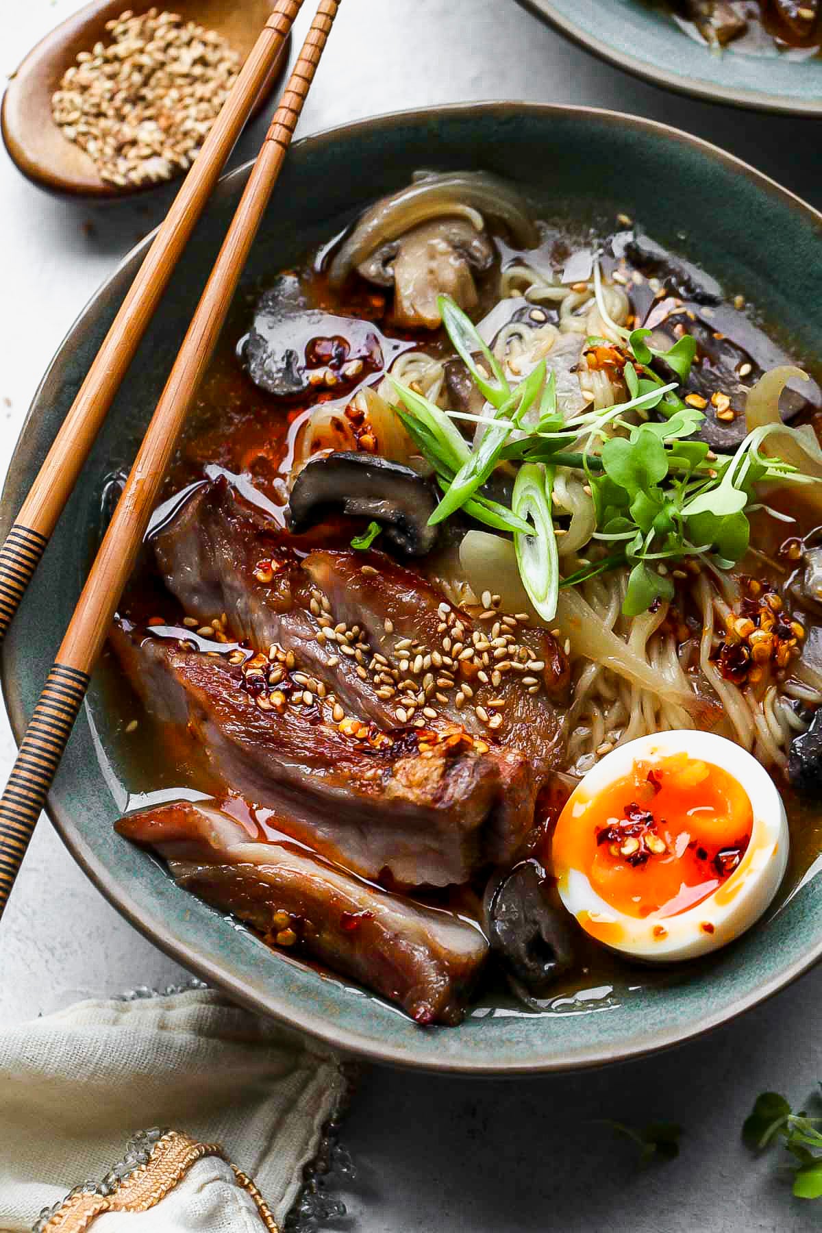 A bowl of assembled slow cooker ramen topped with herbs, egg, and chopsticks resting on the left side.