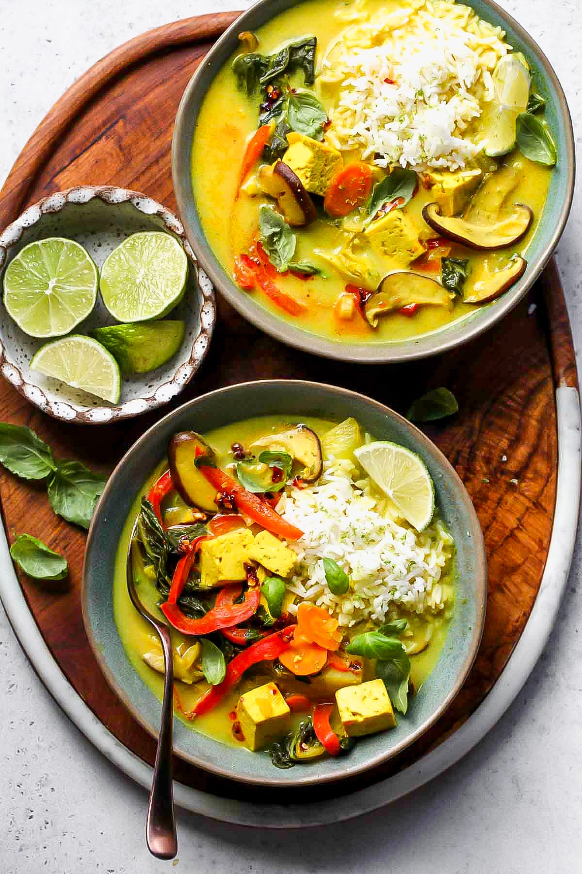 A wooden serving platter with two bowls of thai vegetable soup, as well as a small bowl of lime wedges.