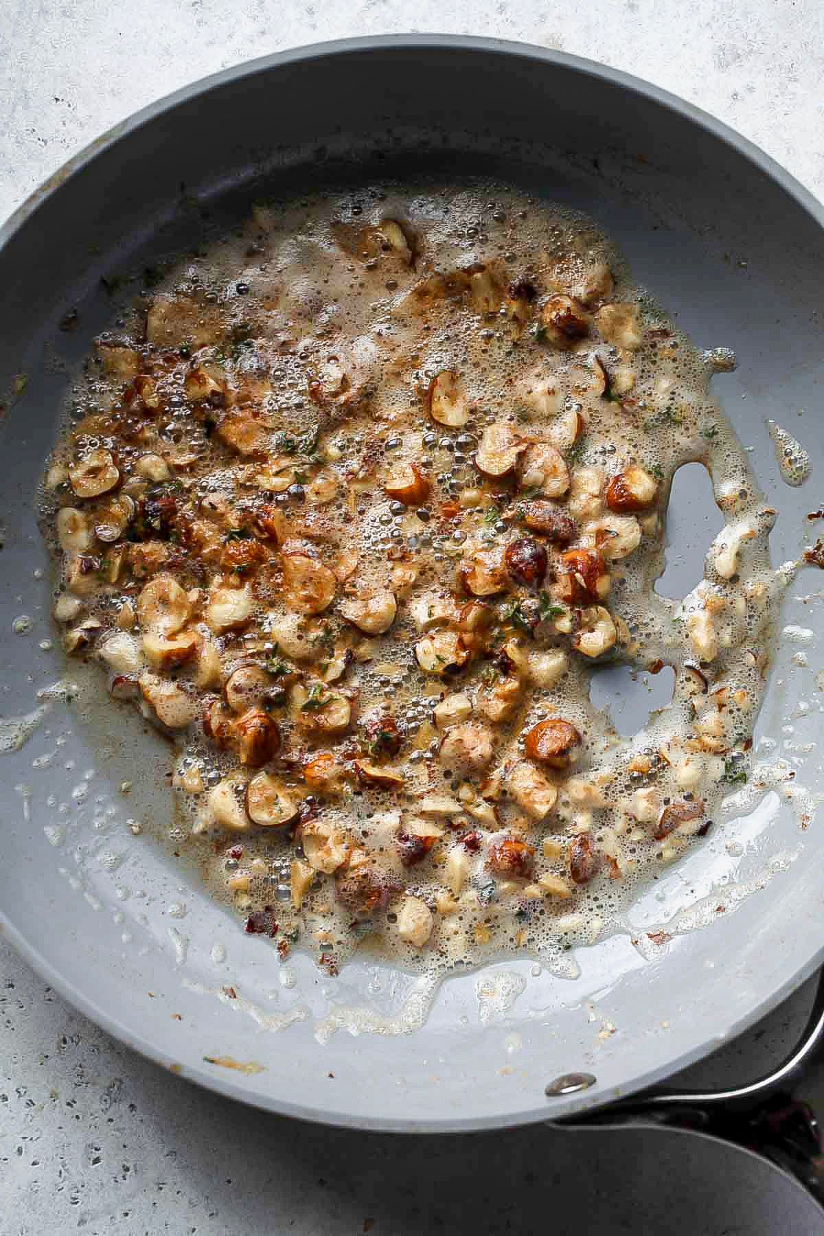 Hazelnuts being cooked in brown butter in a grey skillet.