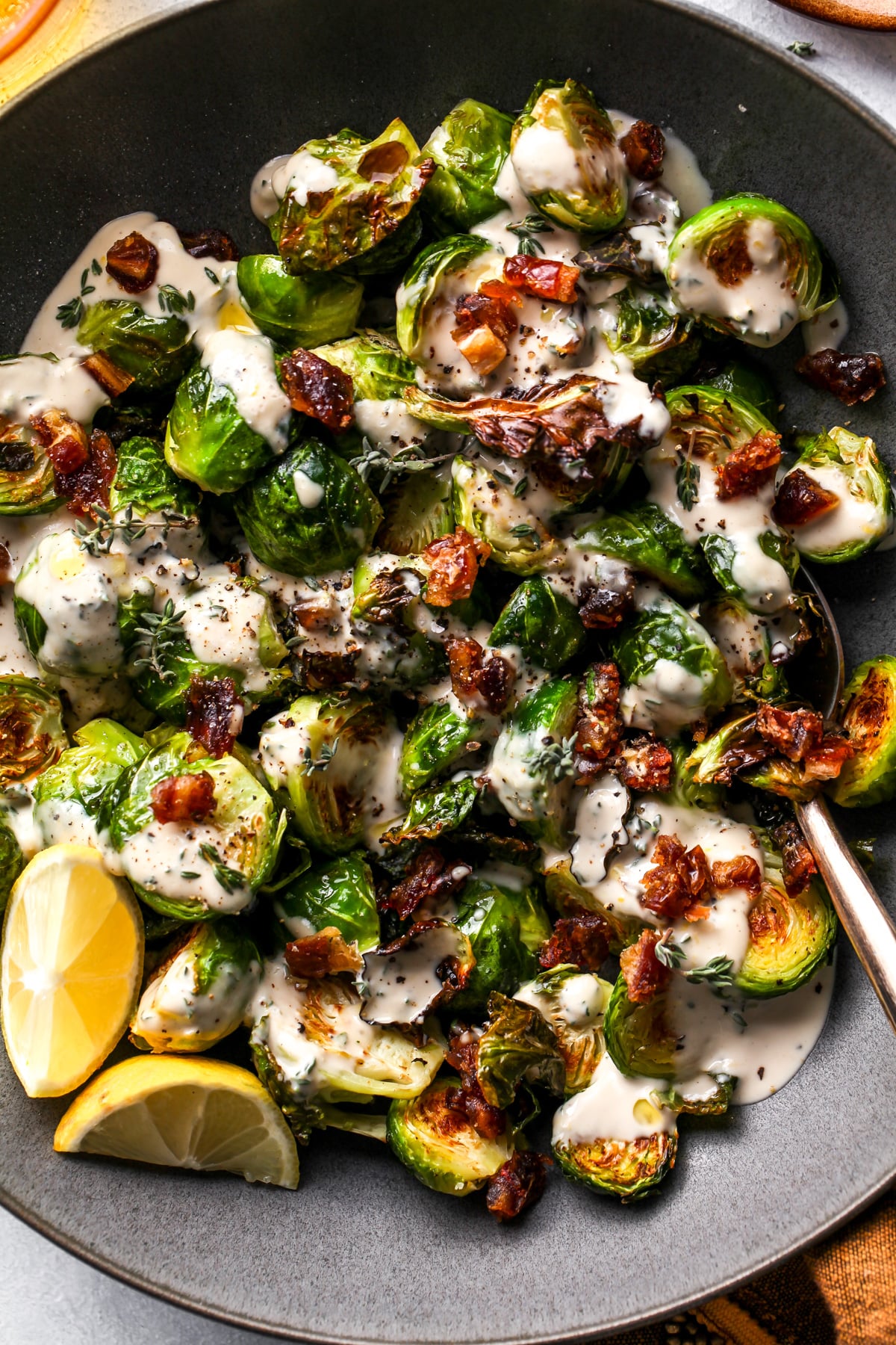 Tahini Brussels sprouts in a dark grey bowl with lemon wedges on the left side.