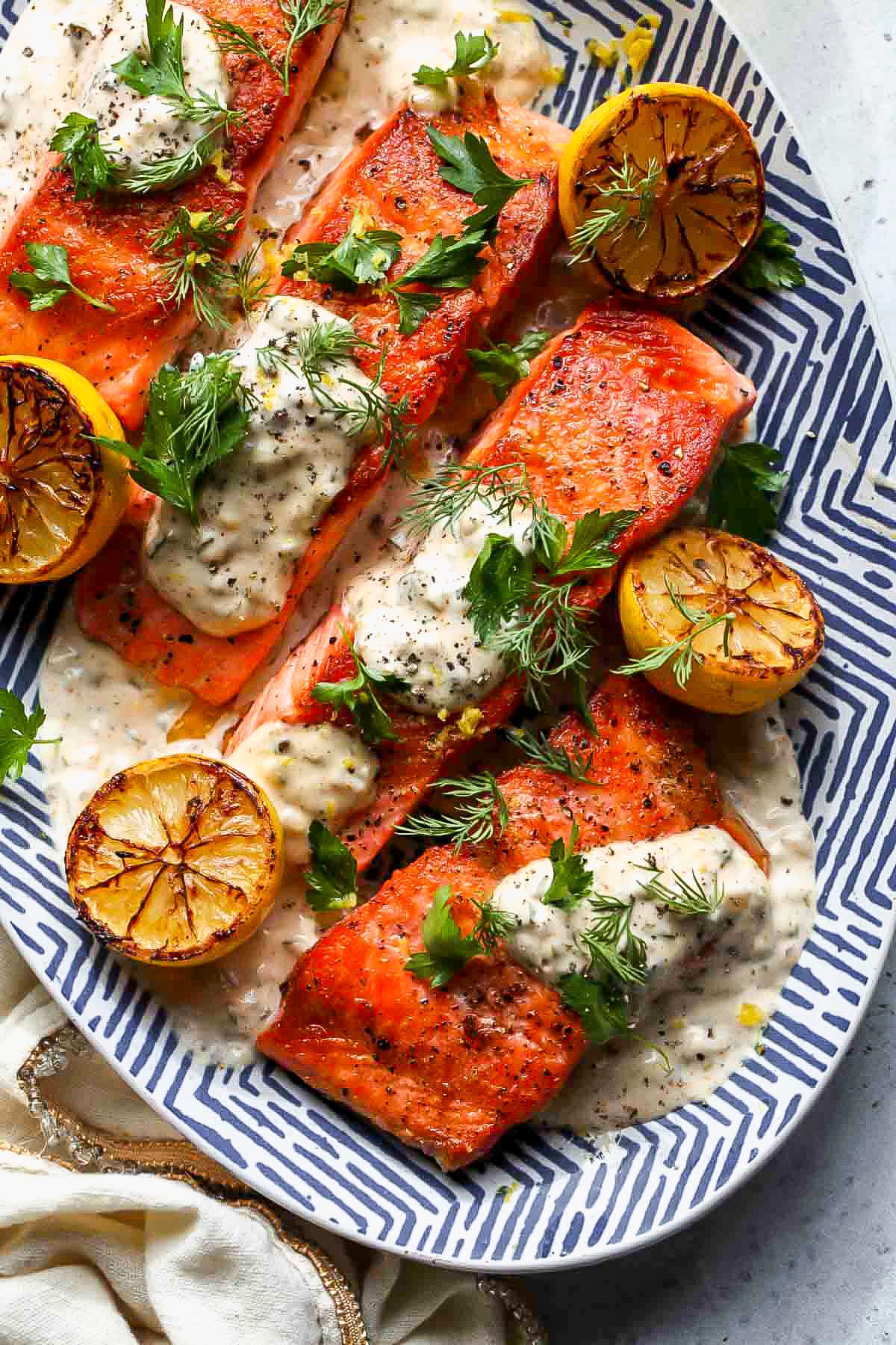 Pan-seared steelhead trout fillets arranged on a blue and white platter with dollops of remoulade, charred lemon halves, and herbs.