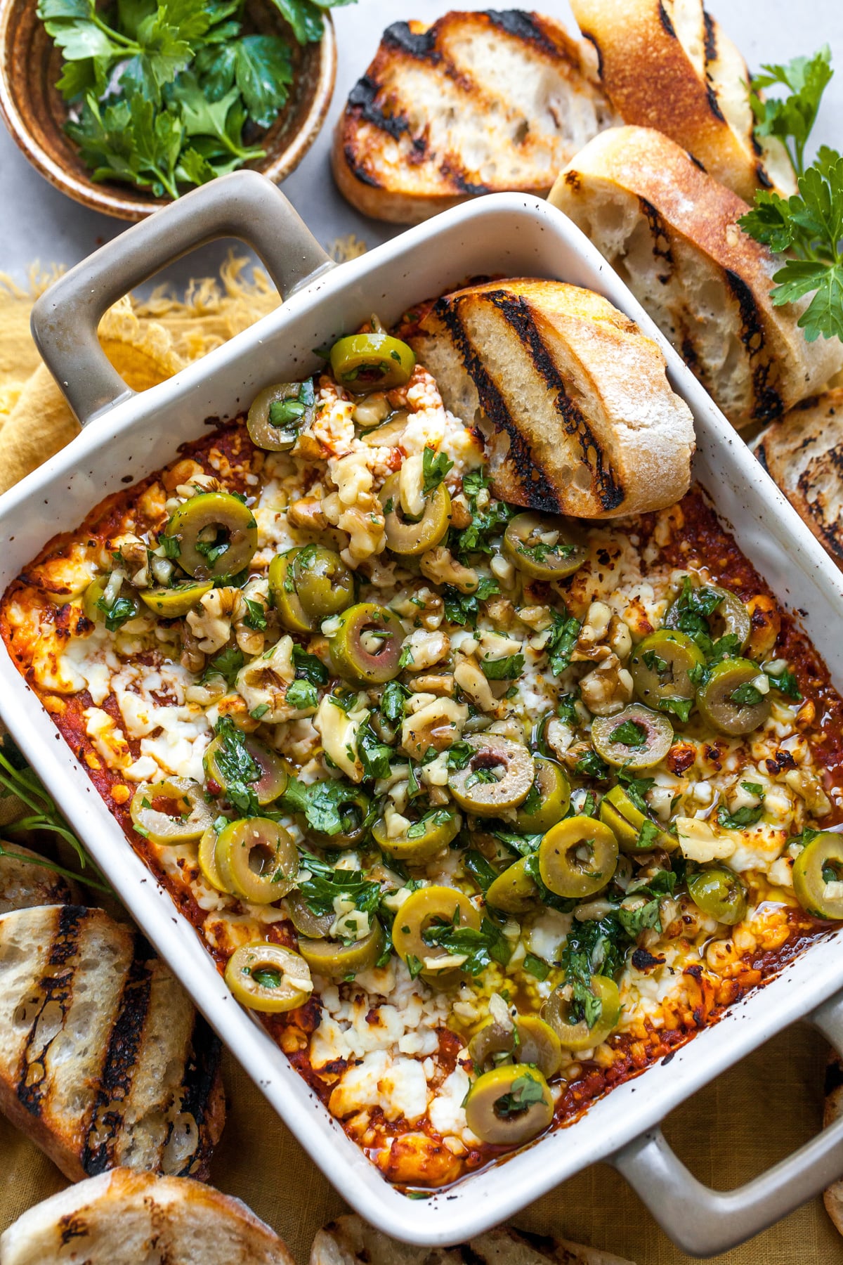 Moroccan Baked Feta dip in a square dish topped with olive tapenade and surrounded by grilled bread.