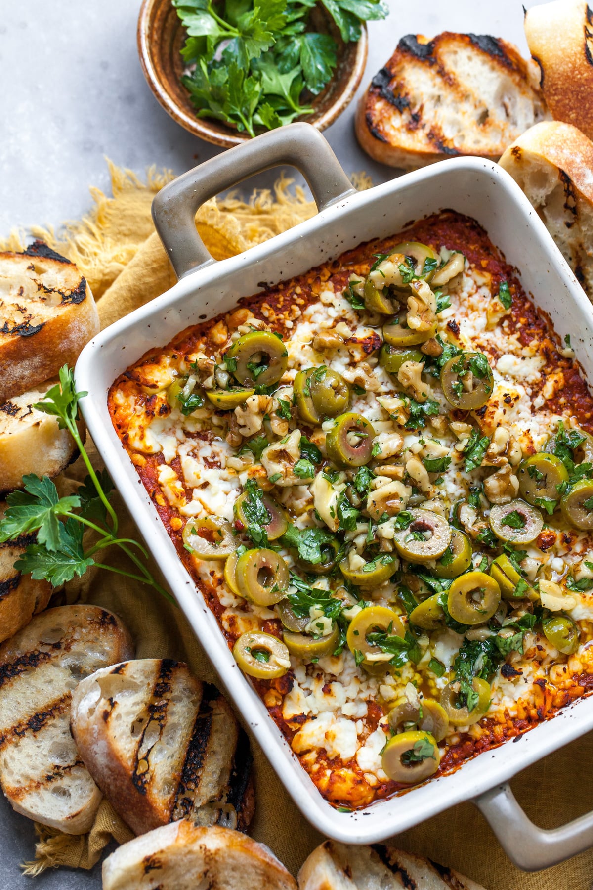Baked feta dip topped with olive tapenade in a square baking dish surrounded by crostini.