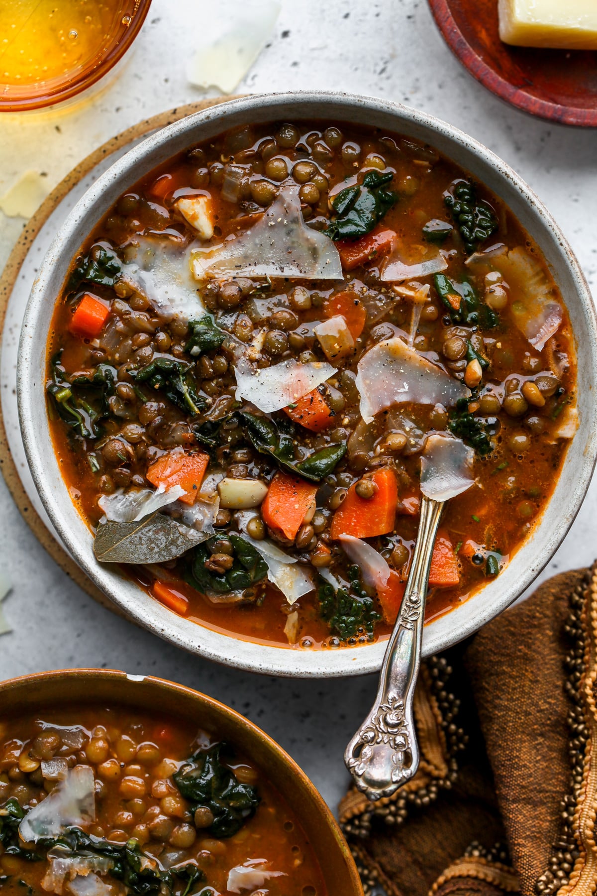 A bowl of lentil soup topped with shaved Parmesan styled on a grey surface with a spoon resting on the right side of the bowl.