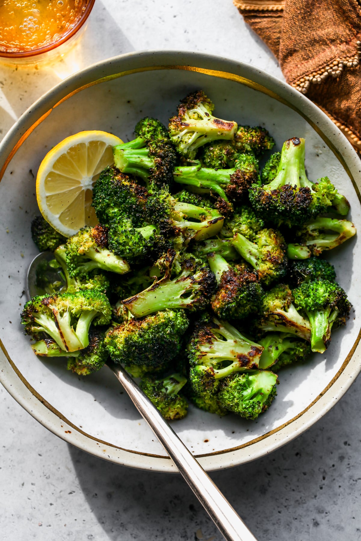 A tan and gold-rimmed bowl filled with sautéed broccoli florets and one wedge of lemon.