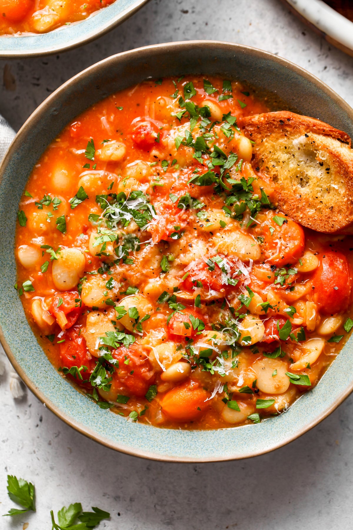 A blue bowl filled with a single portion of tomato and bean stew with a piece of crusty bread on the left side.
