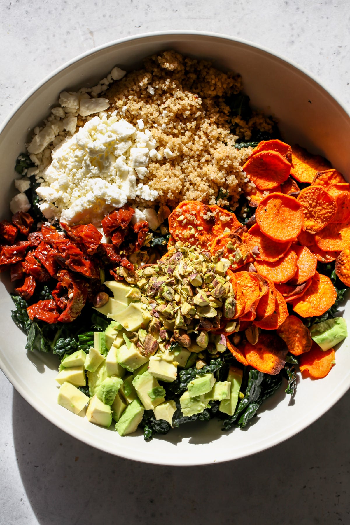 Kale, sweet potato slices, feta, avocado, sun-dried tomatoes, and pistachios in a large white bowl.