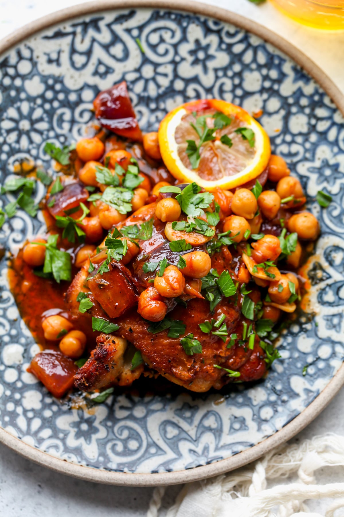 A blue patterned plate with one chicken thigh and a scoop of spiced chickpeas. Garnishes include a lemon slice and chopped parsley.