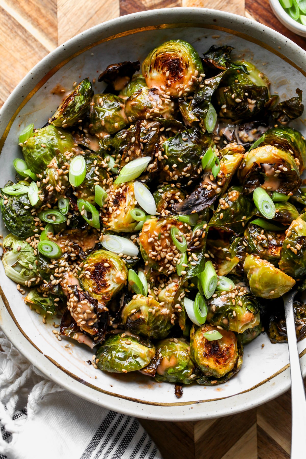Creamy Sriracha Brussels Sprouts styled in a tan and gold trimmed bowl topped with sesame seeds and scallions.