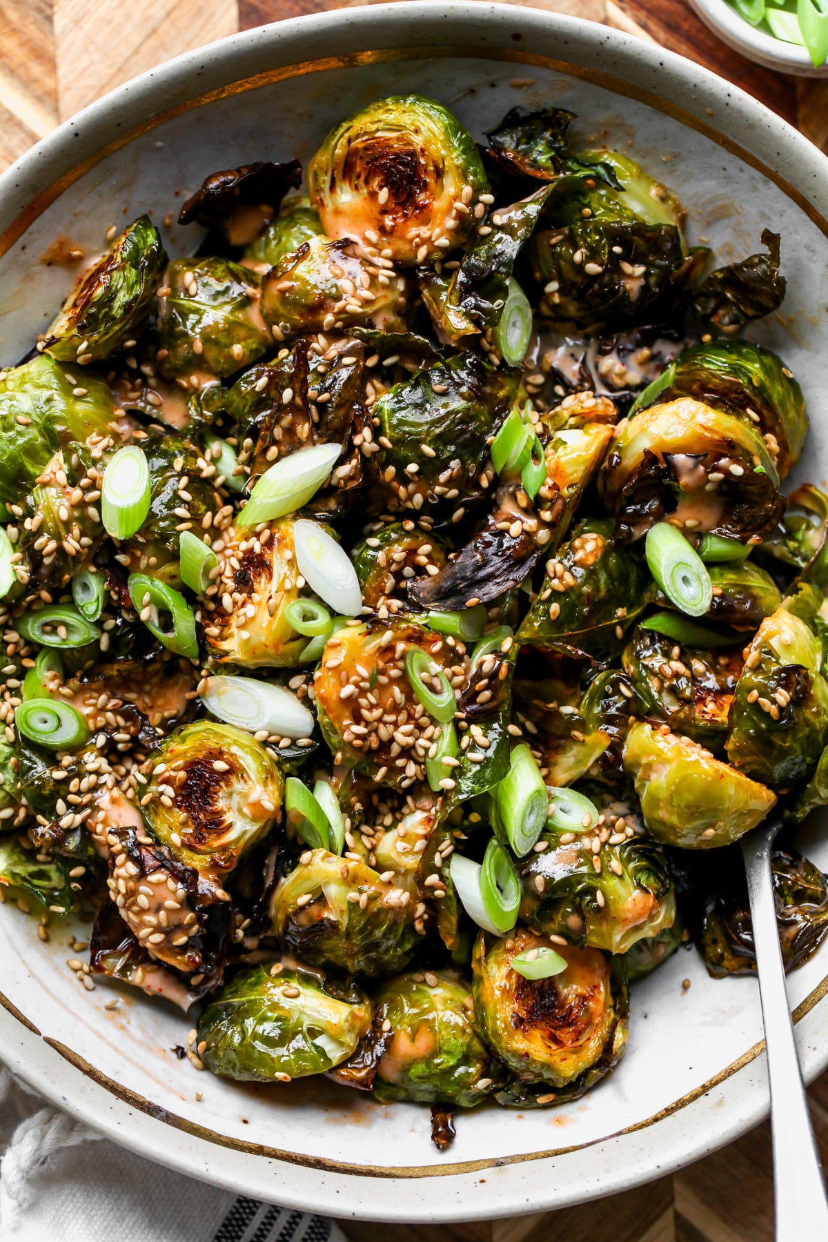 Sriracha brussels sprouts styled in a tan and gold trimmed bowl topped with scallions and sesame seeds. Spoon resting on the right side.