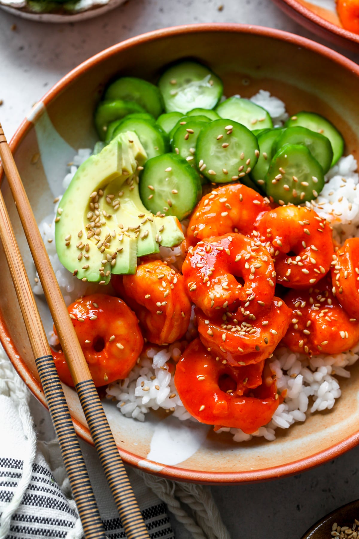 Honey-gochujang shrimp served over rice with cucumbers and sliced avocado in an orange and white serving bowl.