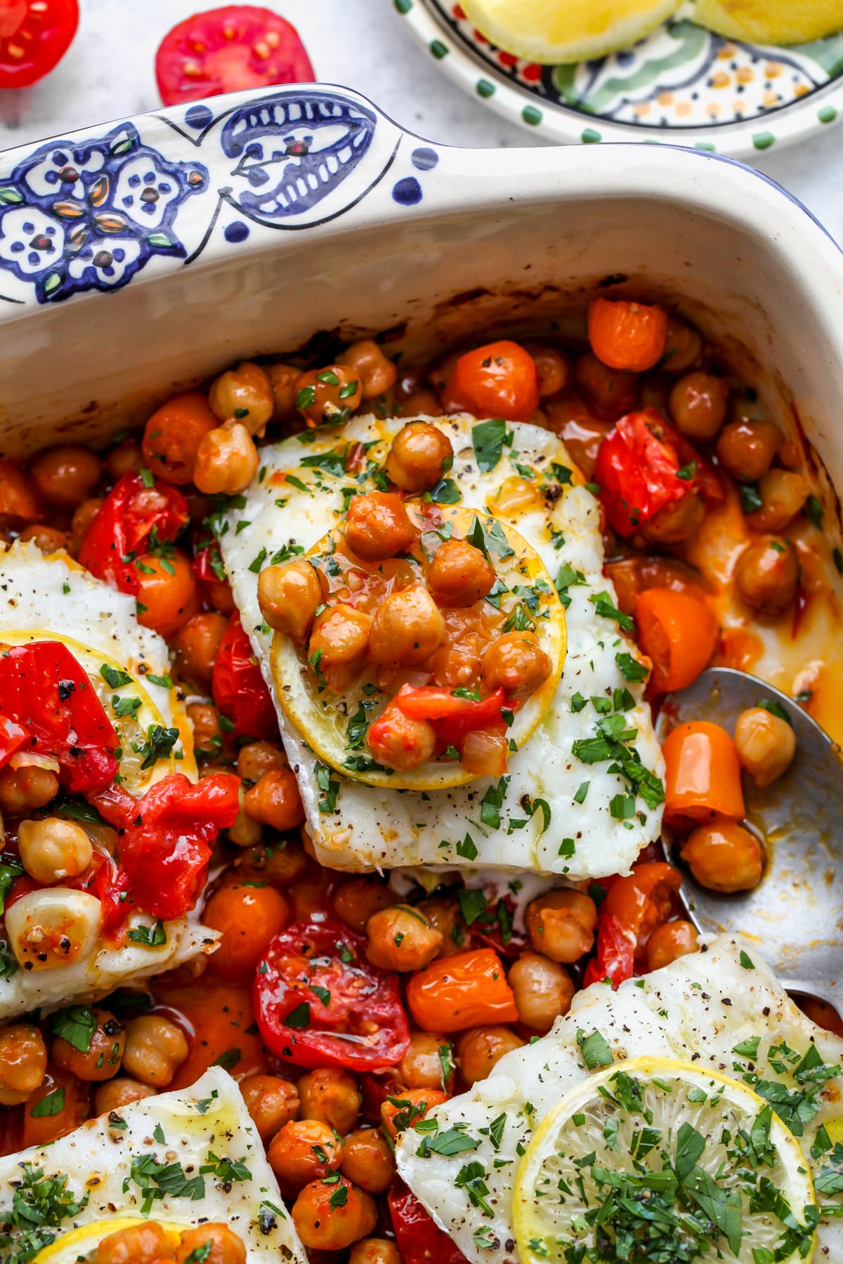 Chickpea and tomato mixture being spooned over cod fillets in a blue and white baking dish.